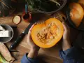 Manos sostienen una calabaza cortada, revelando sus semillas. Ingredientes frescos como queso, cebolleta y especias sugieren la preparación de un plato con semillas de calabaza y sus propiedades.
