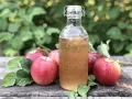 Botella de vinagre de sidra de manzana burbujeante junto a manzanas rojas frescas en una mesa de madera rústica.