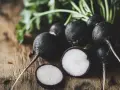 Un grupo de rábanos negros con sus hojas verdes sobre una mesa de madera. Uno está cortado, mostrando su interior blanco y morado.