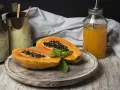 Una papaya fruta cortada por la mitad, con semillas negras y una hoja de menta, junto a un vaso de jugo de papaya.