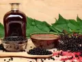 Botella de jarabe oscuro, cuencos con bayas de sauco y hojas verdes.