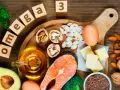 Bloques de madera deletrean "omega 3" junto a salmón, aguacate, nueces, semillas, huevos y aceite, mostrando alimentos ricos en omega 3.