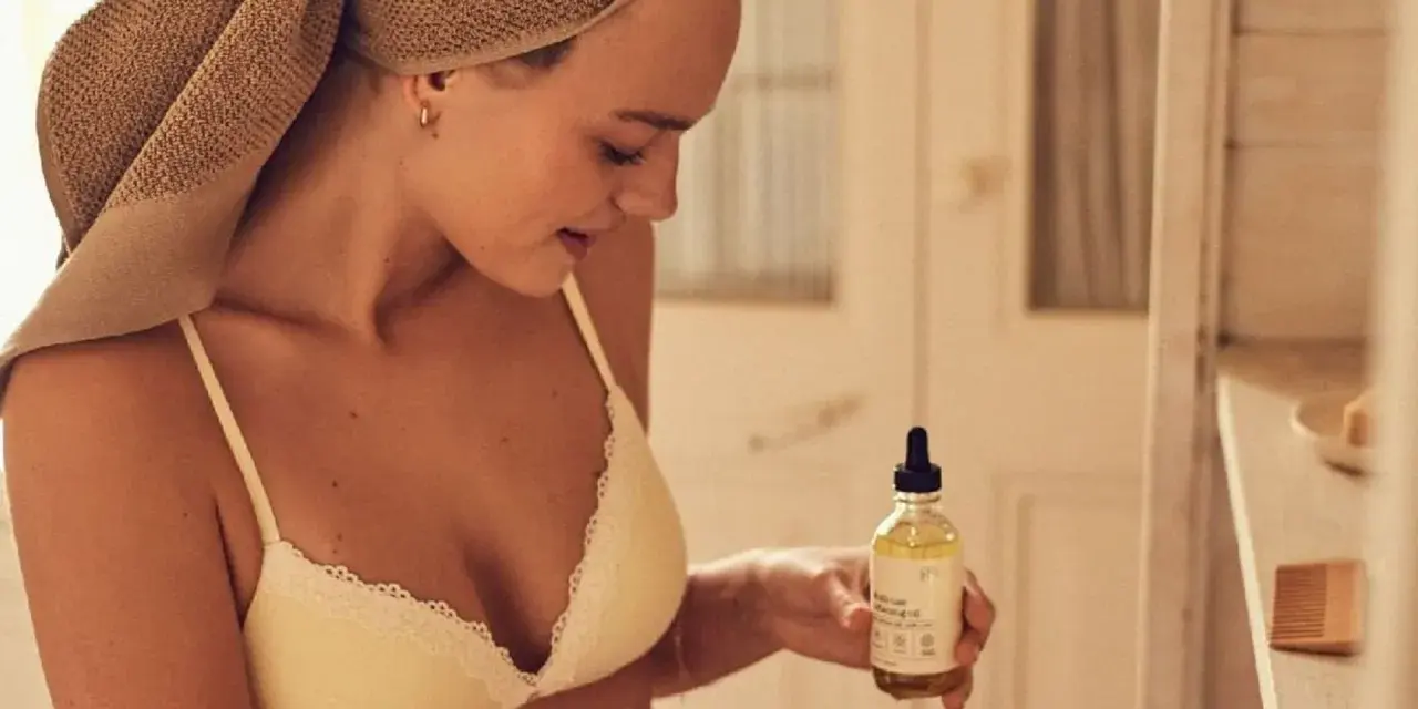 Mujer con toalla en la cabeza sostiene un frasco de aceite de coco, ideal para el cuidado de la piel y el cabello.
