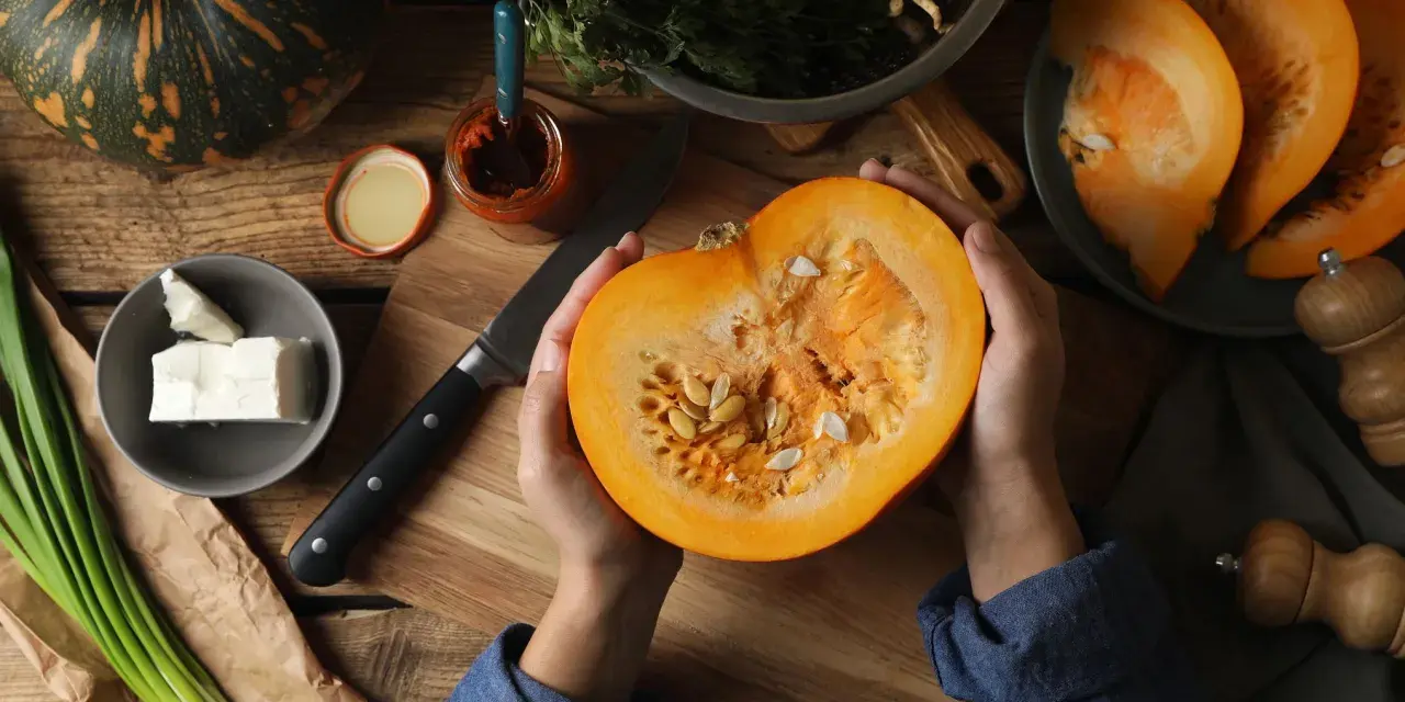 Manos sostienen una calabaza cortada, revelando sus semillas. Ingredientes frescos como queso, cebolleta y especias sugieren la preparación de un plato con semillas de calabaza y sus propiedades.