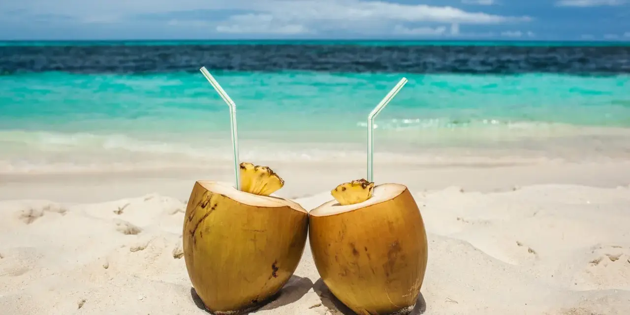 Dos cocos con pajitas en la arena, listos para disfrutar de los agua de coco beneficios. El mar turquesa al fondo.