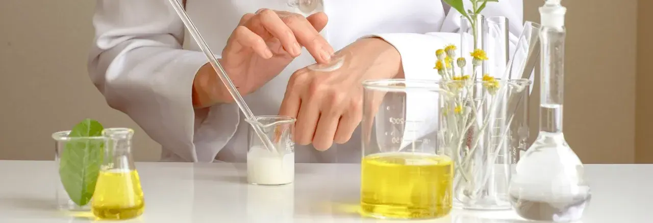 Manos de científico aplicando crema, con tubos de ensayo y líquidos amarillos. El cetearyl alcohol es clave en esta formulación.