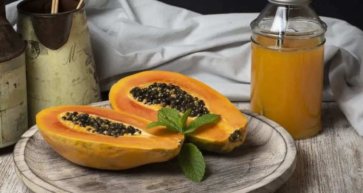 Una papaya fruta cortada por la mitad, con semillas negras y una hoja de menta, junto a un vaso de jugo de papaya.