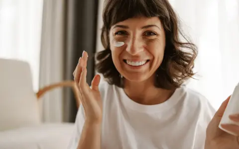 Mujer sonriente aplicándose crema en la mejilla, como si fueran vitaminas para el sol.