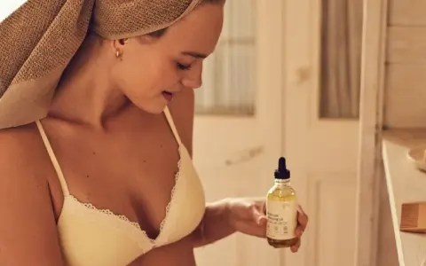 Mujer con toalla en la cabeza sostiene un frasco de aceite de coco, ideal para el cuidado de la piel y el cabello.