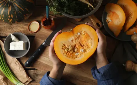 Manos sostienen una calabaza cortada, revelando sus semillas. Ingredientes frescos como queso, cebolleta y especias sugieren la preparación de un plato con semillas de calabaza y sus propiedades.