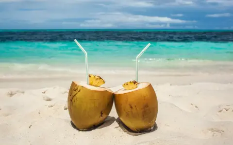 Dos cocos con pajitas en la arena, listos para disfrutar de los agua de coco beneficios. El mar turquesa al fondo.