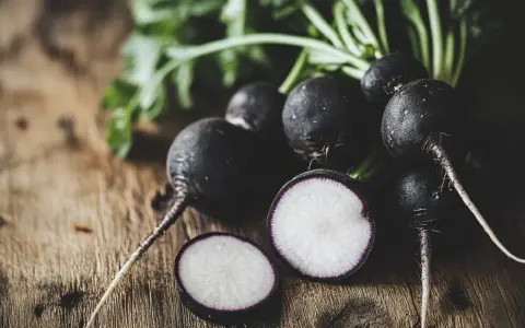 Un grupo de rábanos negros con sus hojas verdes sobre una mesa de madera. Uno está cortado, mostrando su interior blanco y morado.