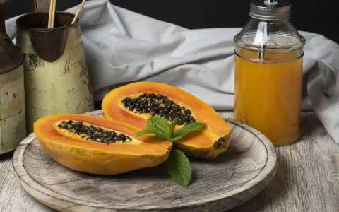 Una papaya fruta cortada por la mitad, con semillas negras y una hoja de menta, junto a un vaso de jugo de papaya.