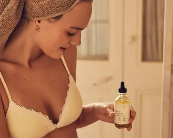 Mujer con toalla en la cabeza sostiene un frasco de aceite de coco, ideal para el cuidado de la piel y el cabello.