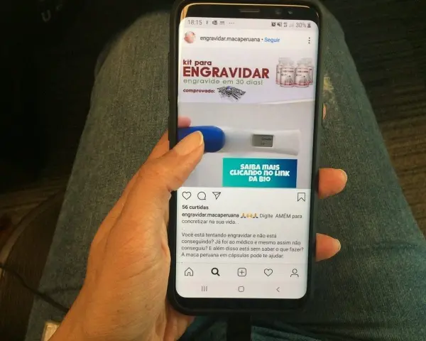 Mano sosteniendo un teléfono que muestra un anuncio de "kit para engravidar" y un test de embarazo. La maca peruana en cápsulas puede ayudar a quienes buscan el embarazo.