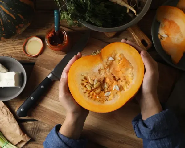 Manos sostienen una calabaza cortada, revelando sus semillas. Ingredientes frescos como queso, cebolleta y especias sugieren la preparación de un plato con semillas de calabaza y sus propiedades.