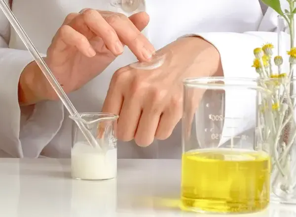 Manos de científico aplicando crema, con tubos de ensayo y líquidos amarillos. El cetearyl alcohol es clave en esta formulación.