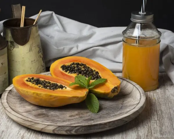 Una papaya fruta cortada por la mitad, con semillas negras y una hoja de menta, junto a un vaso de jugo de papaya.