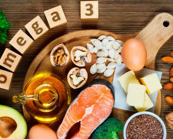 Bloques de madera deletrean "omega 3" junto a salmón, aguacate, nueces, semillas, huevos y aceite, mostrando alimentos ricos en omega 3.