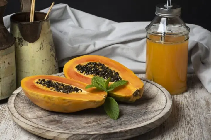 Una papaya fruta cortada por la mitad, con semillas negras y una hoja de menta, junto a un vaso de jugo de papaya.