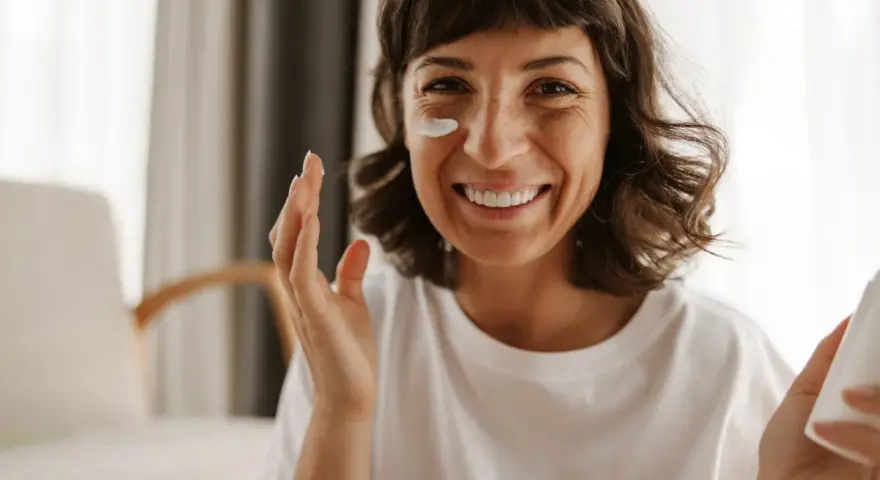 Mujer sonriente aplicándose crema en la mejilla, como si fueran vitaminas para el sol.