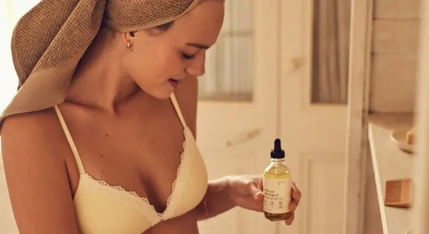 Mujer con toalla en la cabeza sostiene un frasco de aceite de coco, ideal para el cuidado de la piel y el cabello.