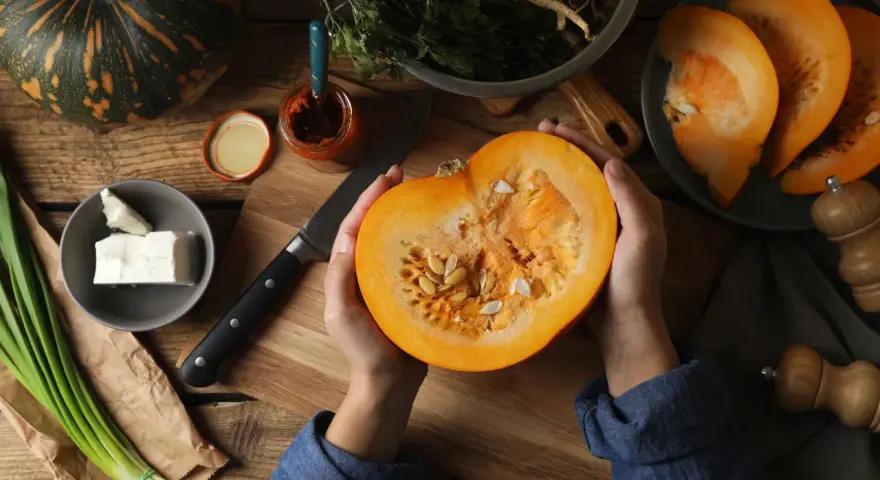 Manos sostienen una calabaza cortada, revelando sus semillas. Ingredientes frescos como queso, cebolleta y especias sugieren la preparación de un plato con semillas de calabaza y sus propiedades.