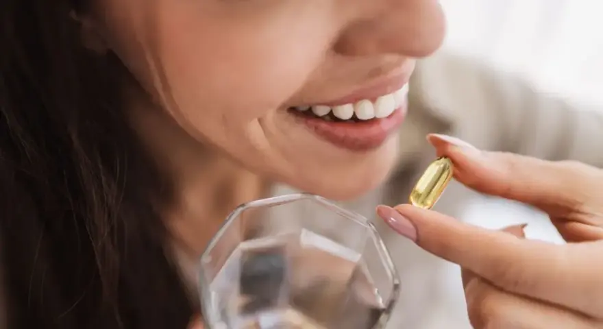 Mujer sonriente toma una cápsula de aceite de pescado con un vaso de agua.