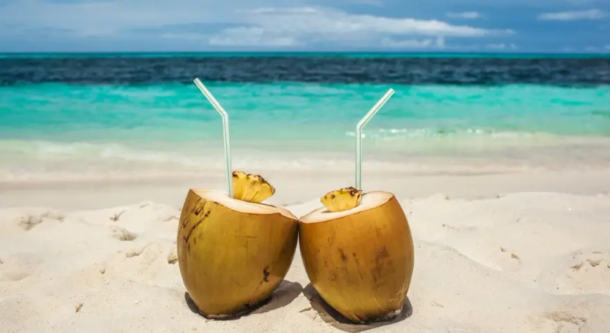 Dos cocos con pajitas en la arena, listos para disfrutar de los agua de coco beneficios. El mar turquesa al fondo.