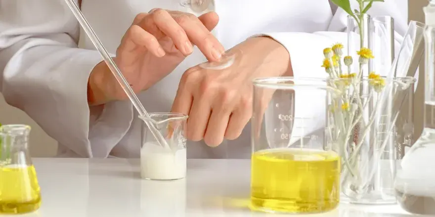 Manos de científico aplicando crema, con tubos de ensayo y líquidos amarillos. El cetearyl alcohol es clave en esta formulación.