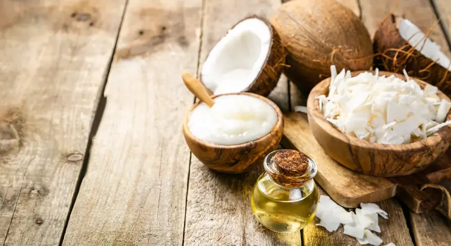 Aceite MCT en una botella junto a cocos partidos, crema de coco y coco rallado sobre madera rústica.