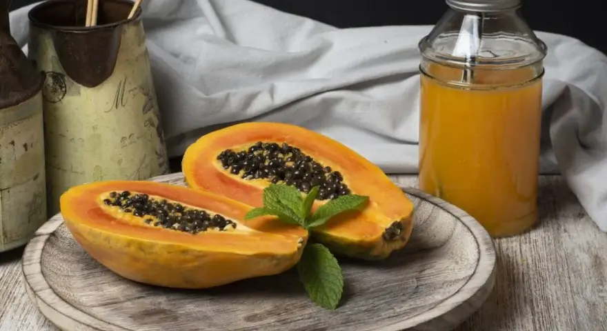 Una papaya fruta cortada por la mitad, con semillas negras y una hoja de menta, junto a un vaso de jugo de papaya.