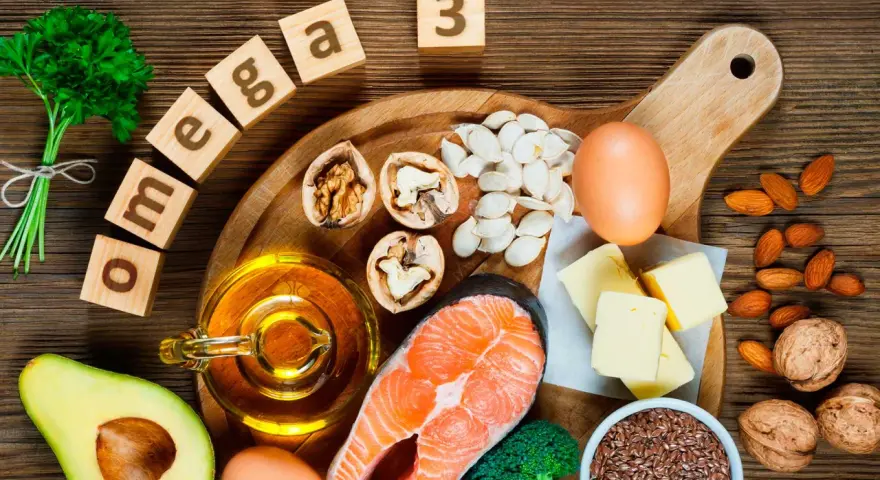Bloques de madera deletrean "omega 3" junto a salmón, aguacate, nueces, semillas, huevos y aceite, mostrando alimentos ricos en omega 3.