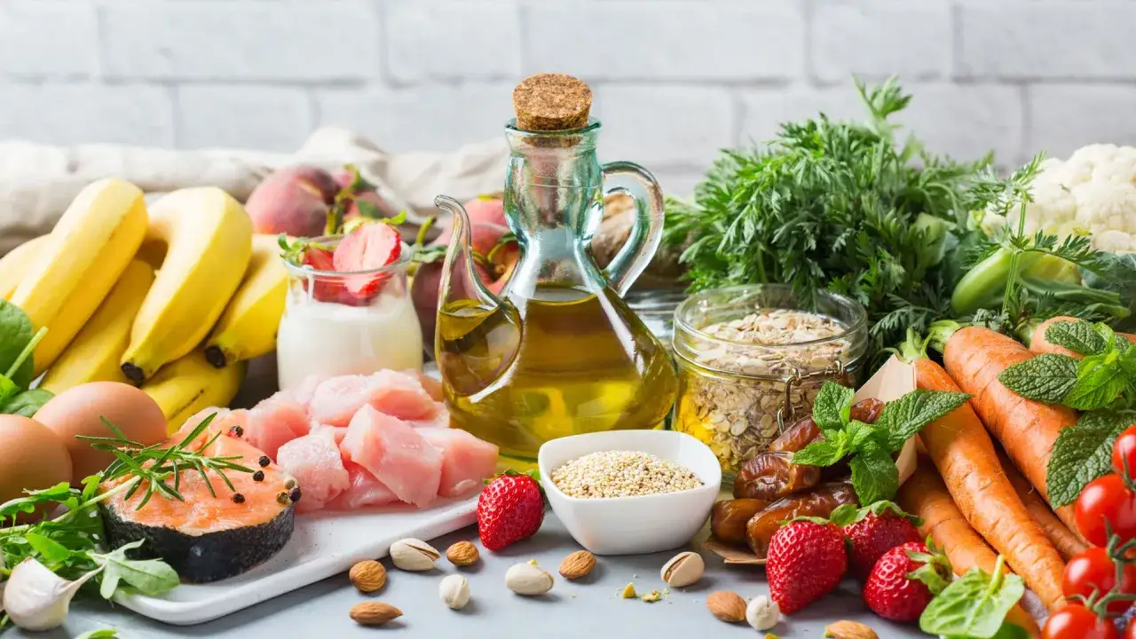 Variedad de alimentos frescos: salm&oacute;n, pollo, huevos, frutas (pl&aacute;tanos, fresas, melocotones), verduras (zanahorias, coliflor, espinacas), frutos secos y aceite de oliva, todos ricos en vitaminas para piel radiante.