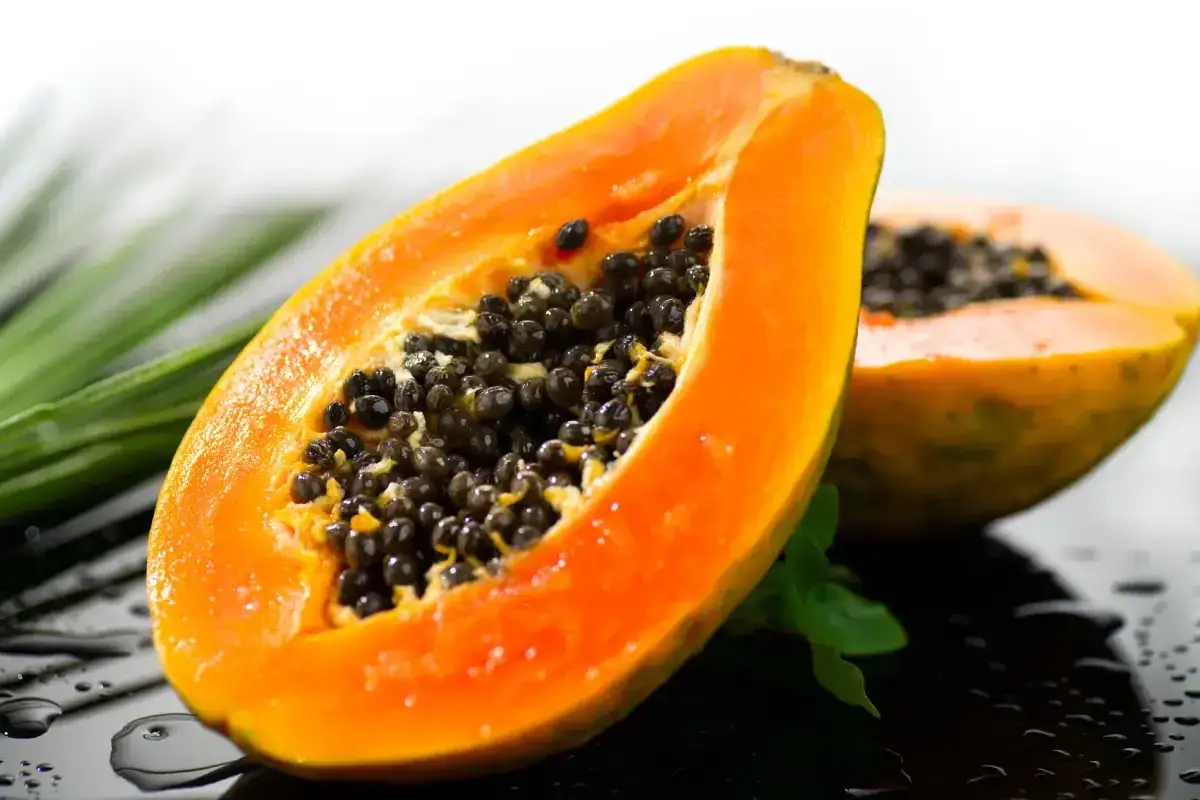 Una jugosa papaya fruta cortada por la mitad, mostrando sus semillas negras y pulpa naranja vibrante, sobre una superficie mojada con hojas verdes.