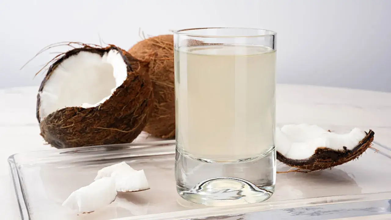 Un vaso de agua de coco fresco junto a cocos partidos. Descubre los agua de coco beneficios para tu salud y bienestar.
